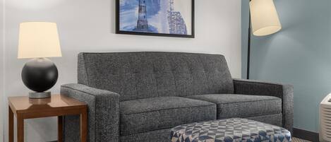 A grey sofa, a wooden side table with a lamp, a patterned ottoman, and a framed picture of a lighthouse on the wall.