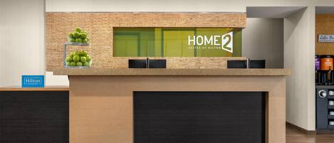 Una recepción con un letrero que dice 'HOME2', un letrero de 'Hilton Honors' y una barra de café al fondo.