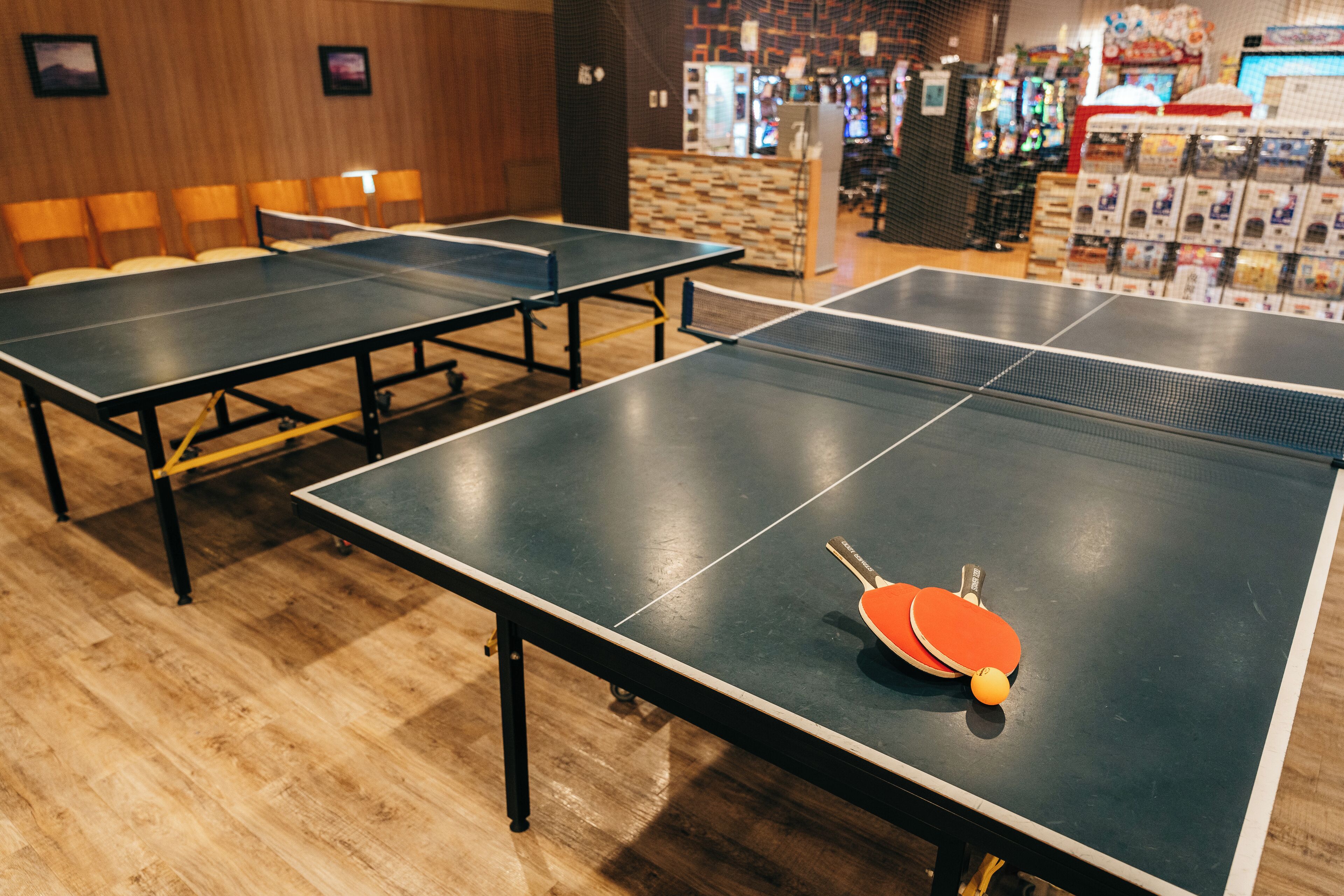 A table tennis table with two paddles and a ball, surrounded by chairs, in a room with wooden flooring and a brick wall.