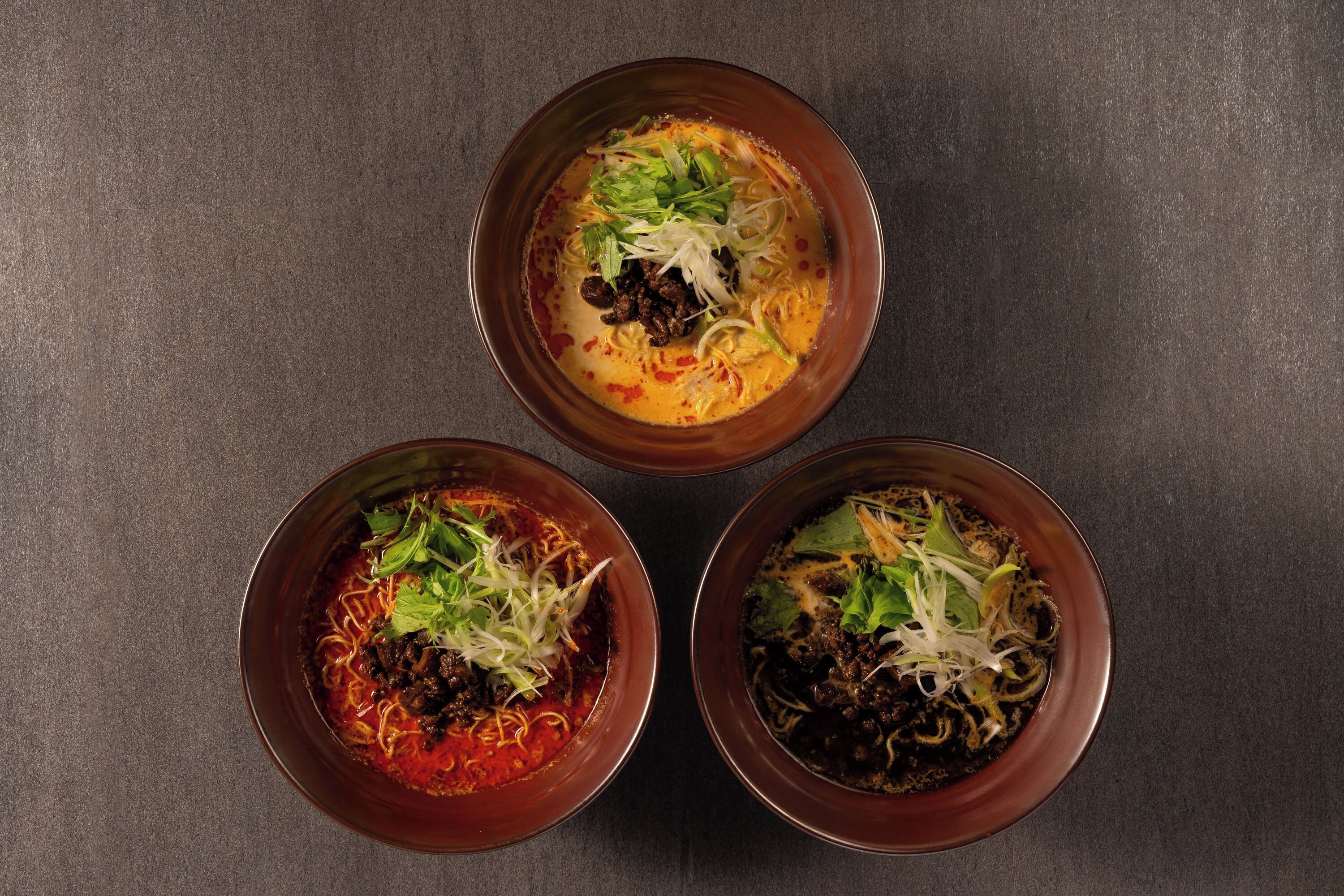Three bowls of soup with noodles and vegetables on a dark surface.
