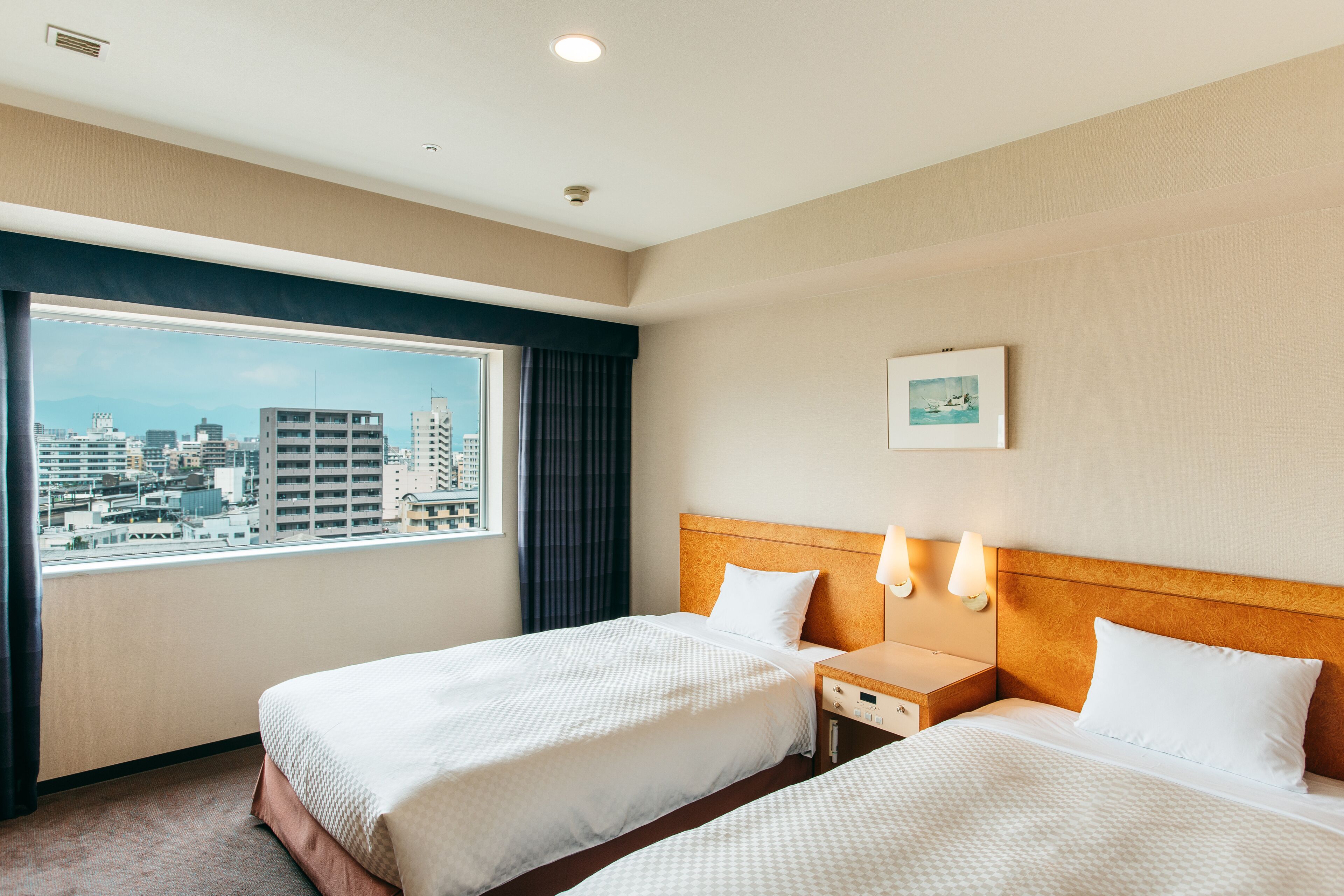 A hotel room with two beds, a large window with a city view, a headboard, bedside lamps, and a small bedside table.