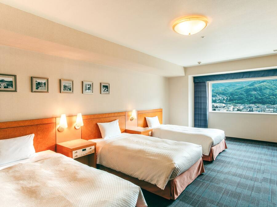 Habitación de hotel con tres camas, un ventanal amplio y vistas a la ciudad, y luces fijadas a la pared.