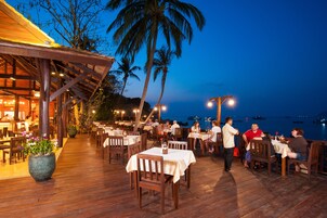 Un restaurante al aire libre con mesas y sillas de madera, palmeras y vistas al mar.