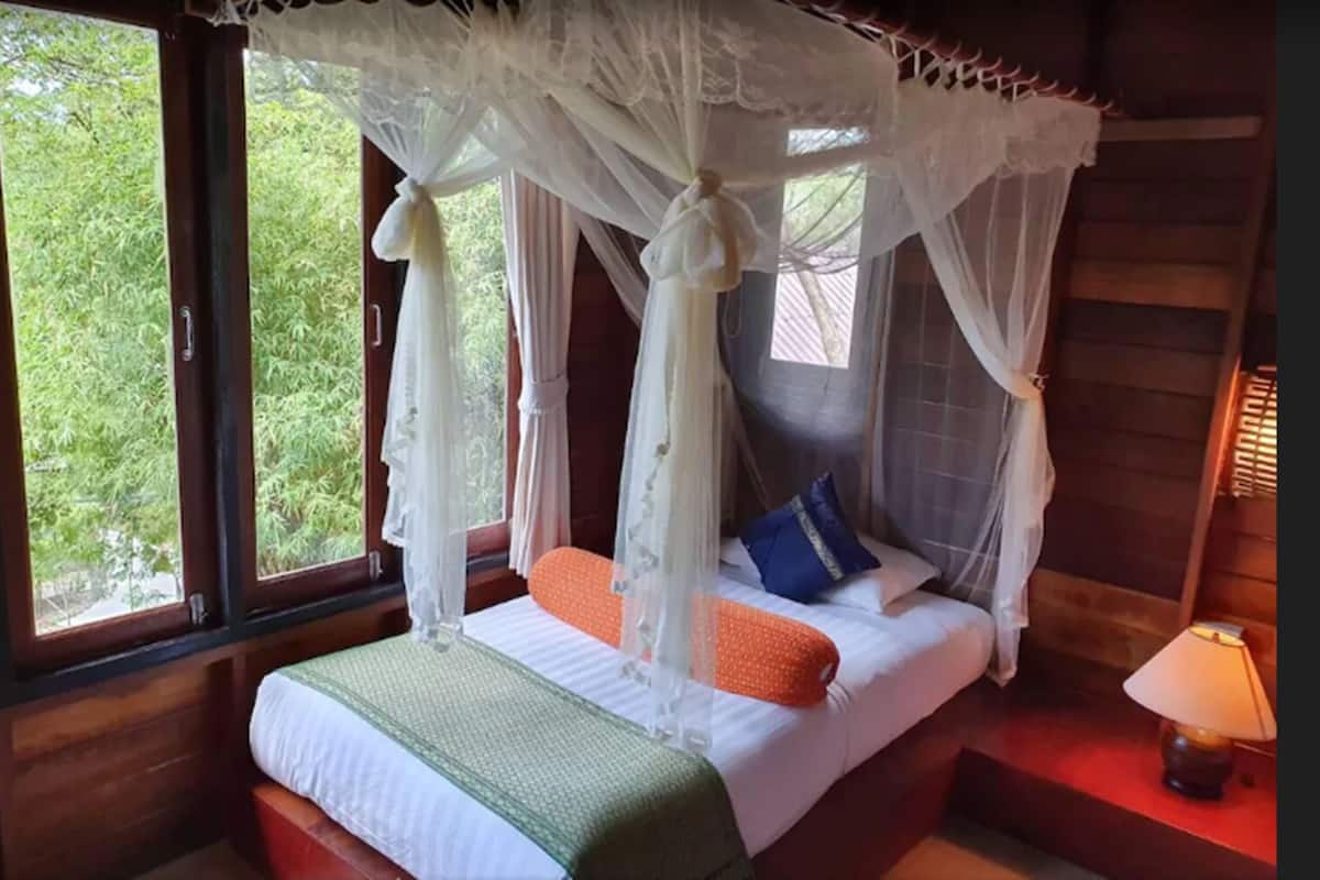A bedroom with a four-poster canopy bed, a bedside table with a lamp, and a view of greenery outside.