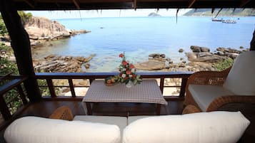 Una terraza de madera con mobiliario de mimbre que da a un litoral rocoso y un mar tranquilo.