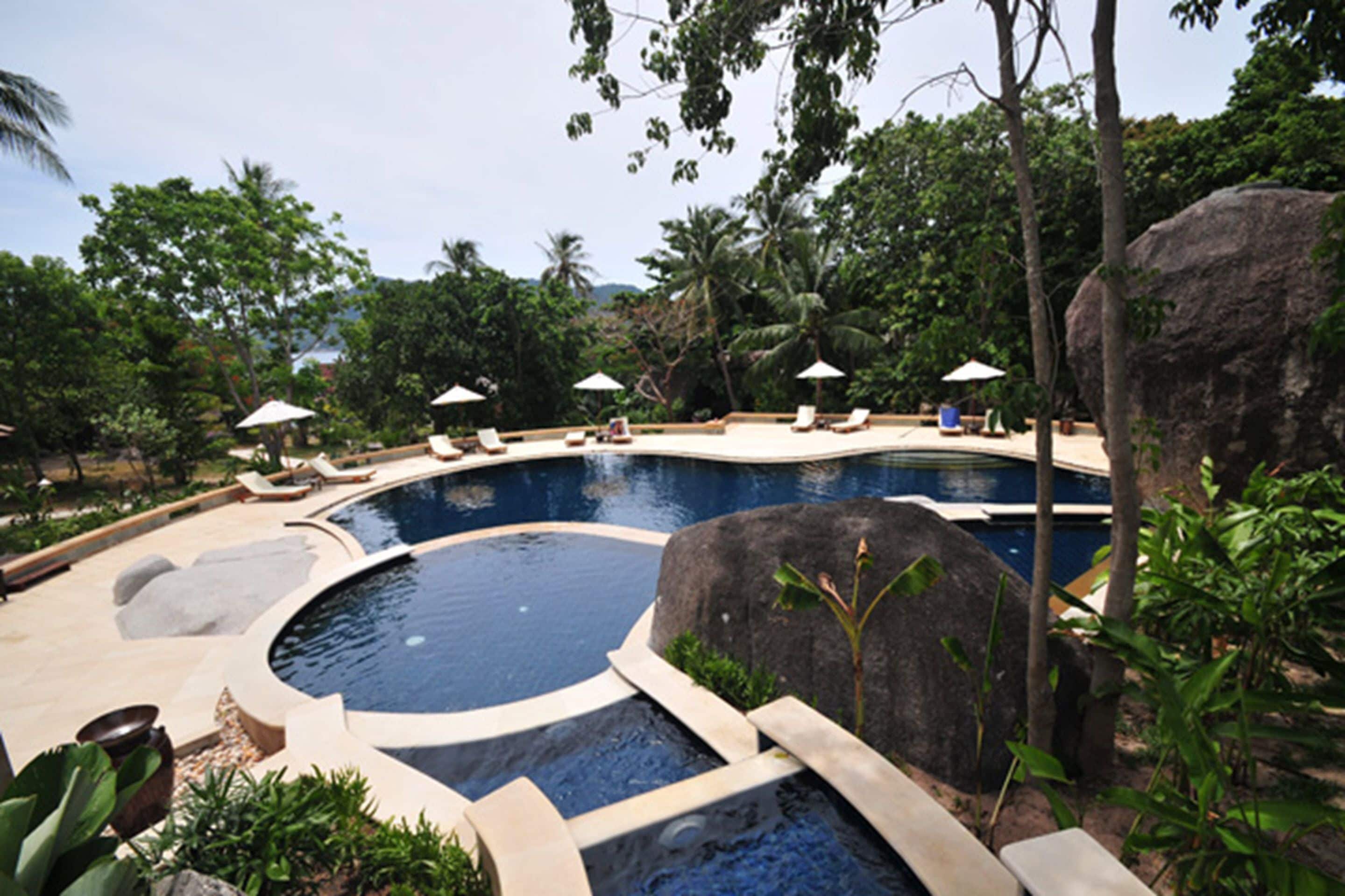 Una piscina con sillas de descanso y sombrillas, rodeada de vegetación tropical y rocas.