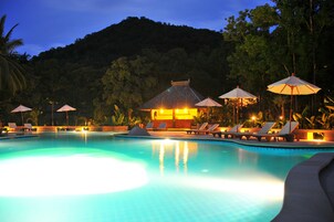 Una piscina con sillones de descanso y sombrillas, rodeada de follaje y una colina al fondo.