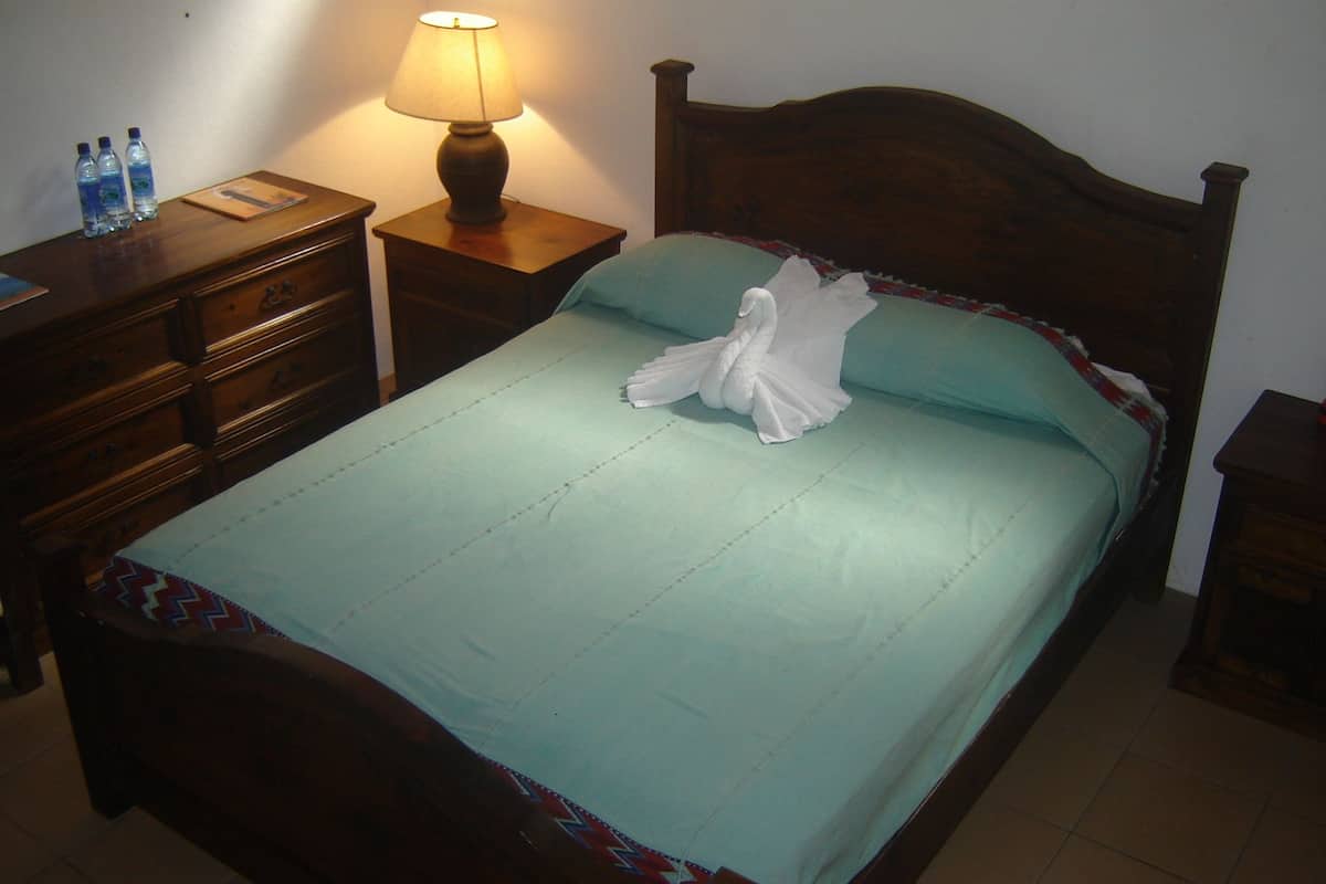 A neatly made bed with a teal bedspread, a white towel folded on the bed, and a wooden headboard. There is a wooden nightstand with a lamp and a few water bottles on it.