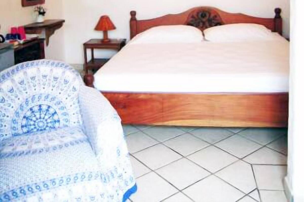 Ein Schlafzimmer mit einem hölzernen Bett, Deckenventilator, Nachttisch, Lampe und einem Stuhl mit gemustertem Bezug.