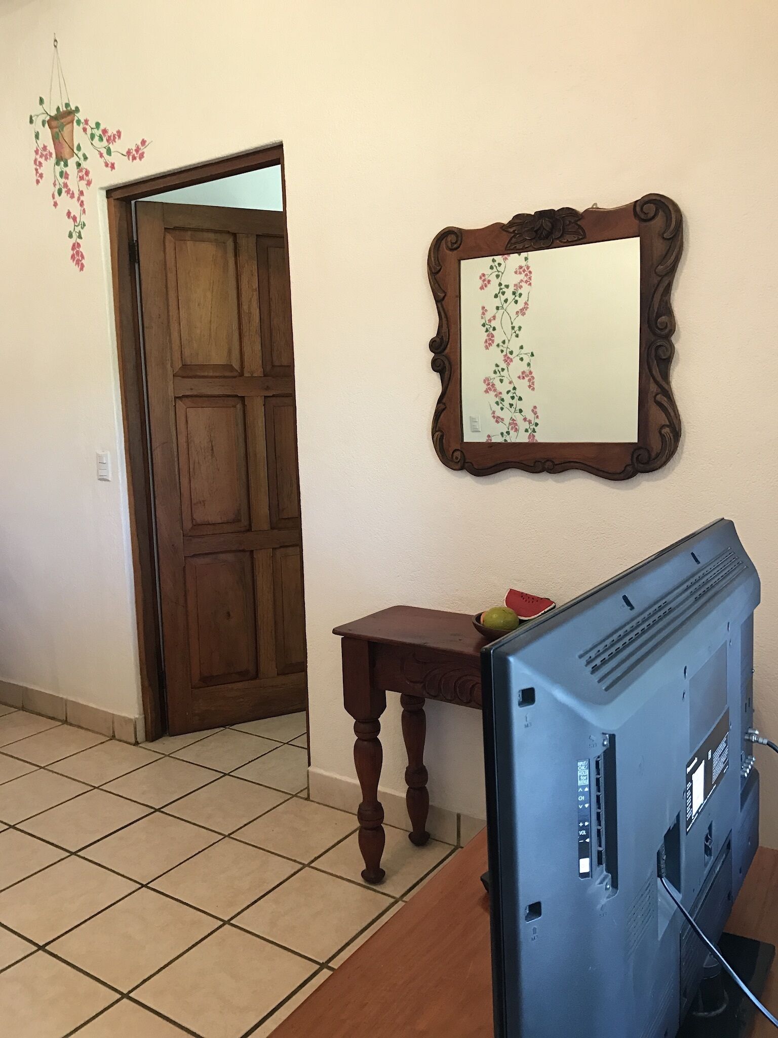 Una habitación con puerta de madera, una pequeña mesa de madera con un cuenco y fruta, un espejo con marco floral, y un televisor sobre un mueble.
