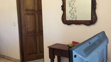 Una habitación con puerta de madera, una pequeña mesa de madera con un cuenco y fruta, un espejo con marco floral, y un televisor sobre un mueble.
