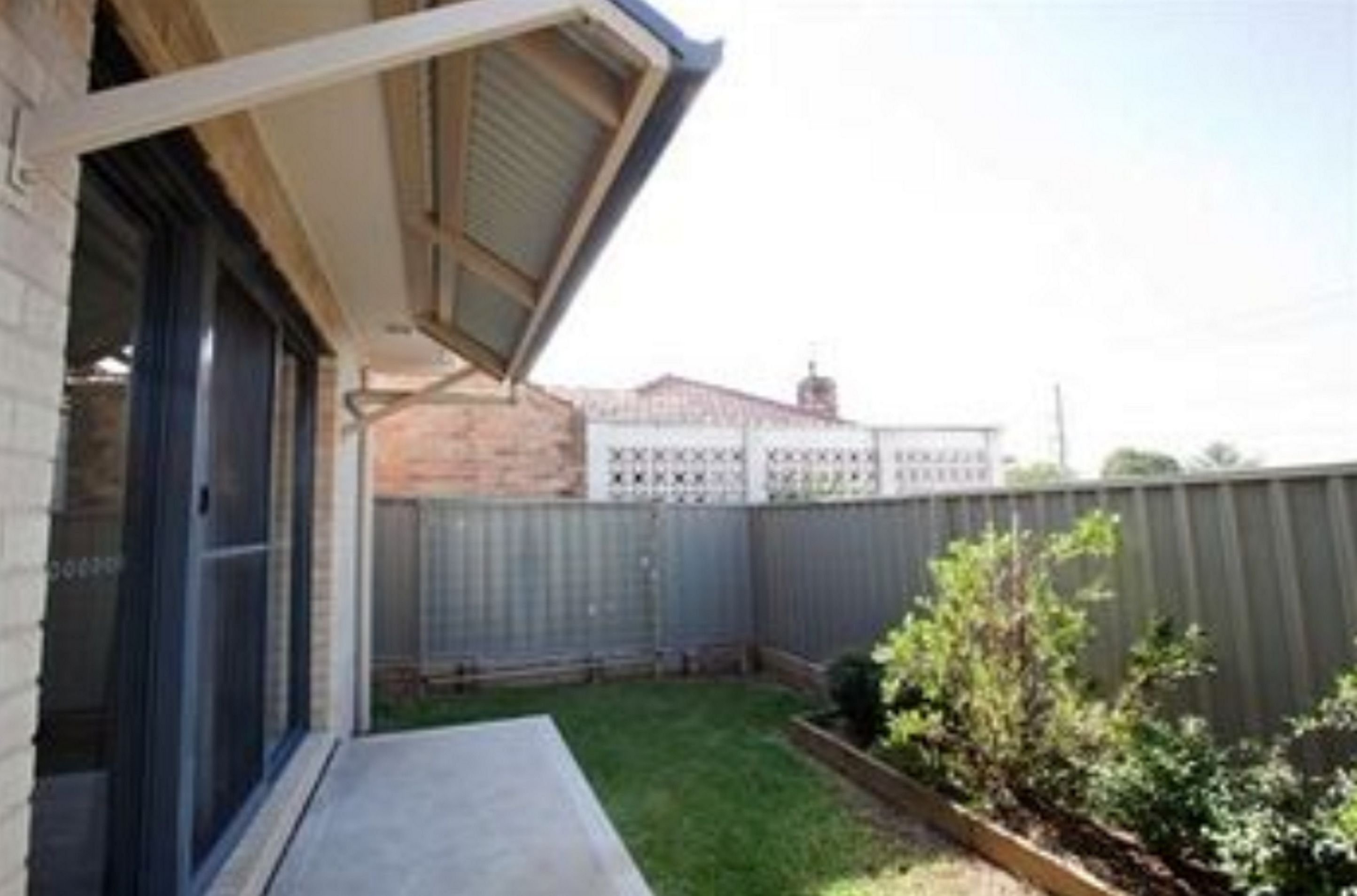 Un patio trasero residencial con puerta corrediza de vidrio, un patio de concreto, una terraza de madera y una cerca de metal.