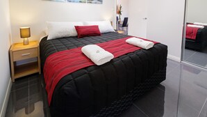 Una habitación de hotel moderna con una cama acolchada negra, almohadas blancas y rojas, una colcha a rayas rojas y negras, y dos toallas dobladas con cuidado.