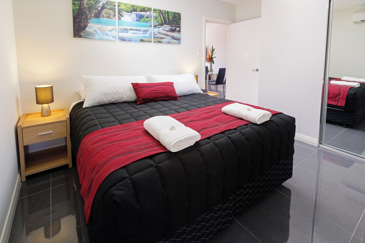 A modern hotel room with a large bed, a bedside table with a lamp, a chair, and a desk.