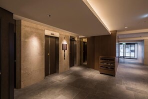 A modern elevator area with three elevators, a wall-mounted sign, and a wooden bench. - Hotel Mystays Nagoya Nishiki (Nagoya)