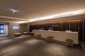 A hotel reception area with a modern design, featuring a reception desk, two staff members, and a large screen displaying information. - Hotel Mystays Nagoya Nishiki (Nagoya)