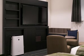 A hotel room with a flat-screen TV mounted on a black wall, a compact air conditioning unit, a small table with a blue pillow, and a brown chair. - Hotel Mystays Nagoya Nishiki (Nagoya)