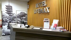 Un comptoir d’accueil d’hôtel avec un mur recouvert de panneaux en bois, une grande enseigne indiquant « HÔTEL LEMAX », ainsi que divers documents d’information et un écran numérique.