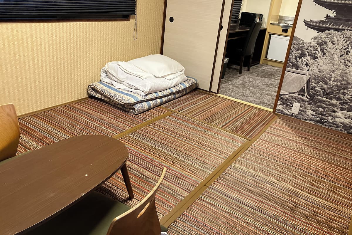 Une chambre d’hôtel avec un sol recouvert de tatamis, une petite table basse avec une chaise, un lit avec une couverture pliée et une porte coulissante donnant sur un bureau équipé d’un ordinateur.