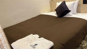 Un lit bien fait, avec une couverture marron, des oreillers blancs et une serviette blanche pliée, ornée d’un motif.