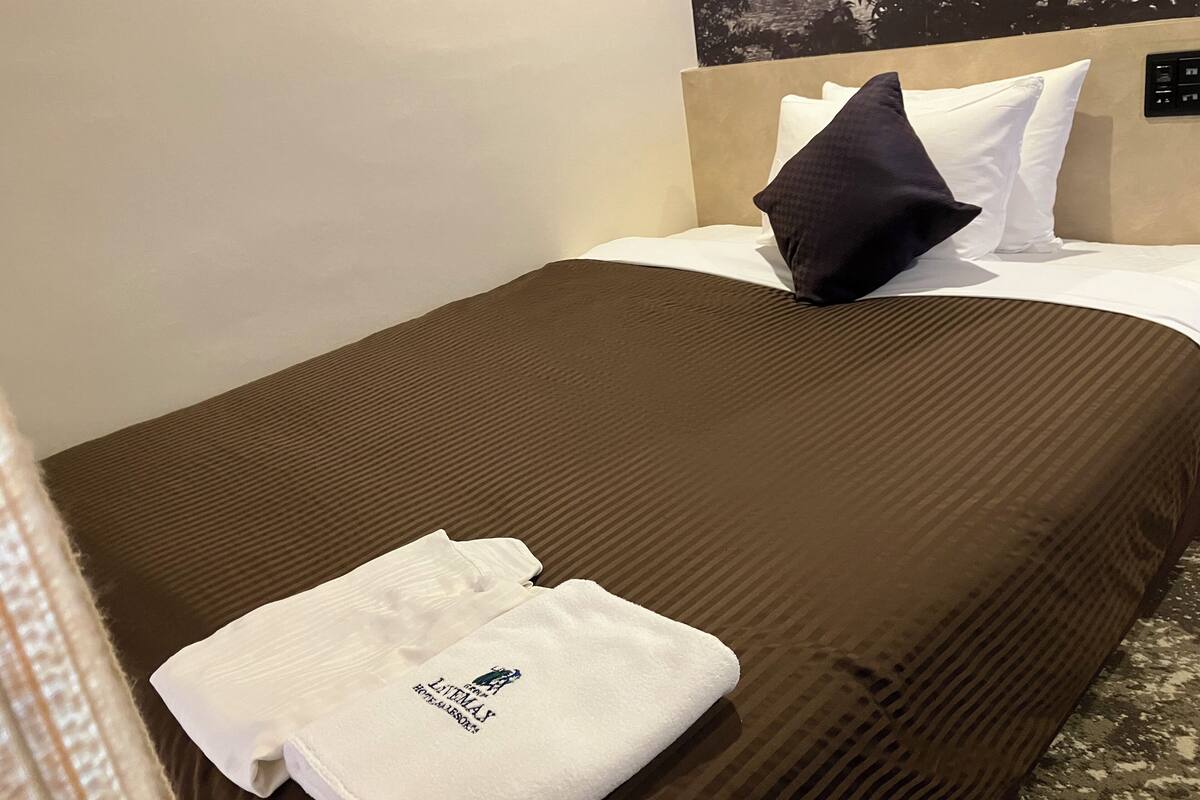 Un lit bien fait, avec une couverture marron, des oreillers blancs et une serviette blanche pliée, ornée d’un motif.