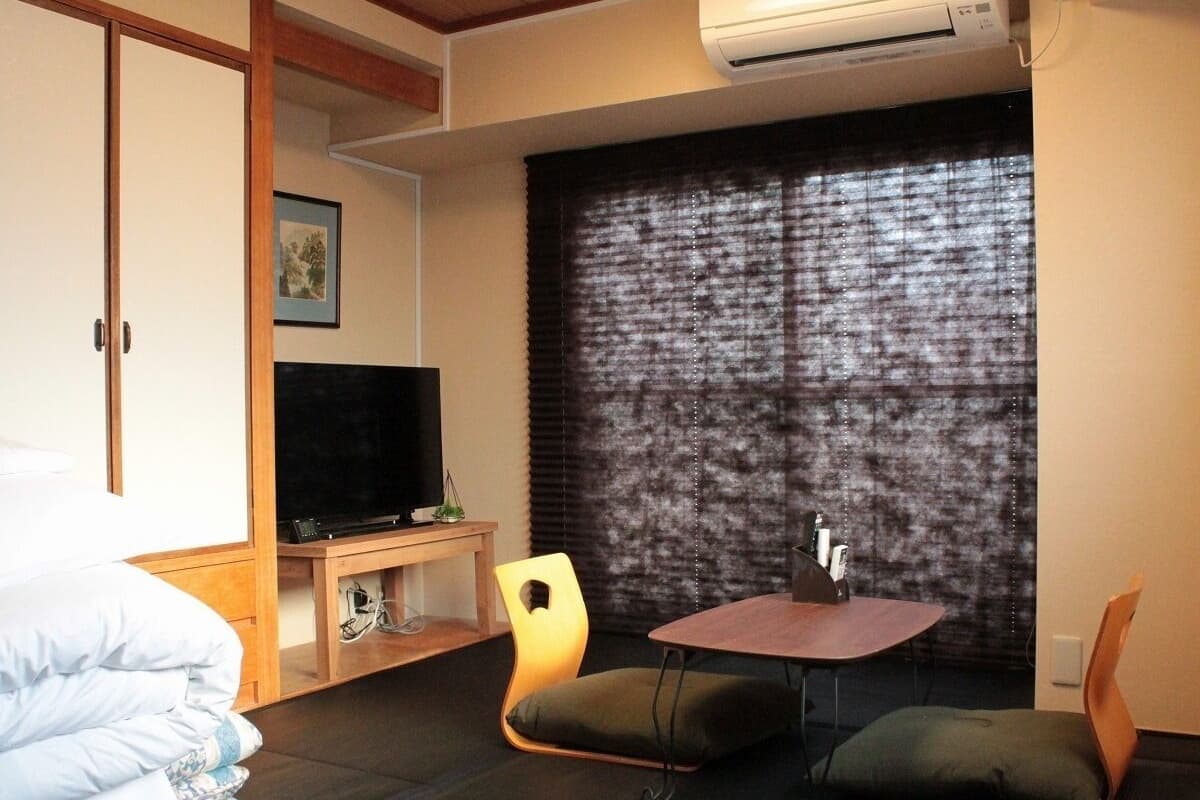 Une chambre d’hôtel avec un lit, une télévision, une petite table avec des chaises et une grande fenêtre avec des stores.