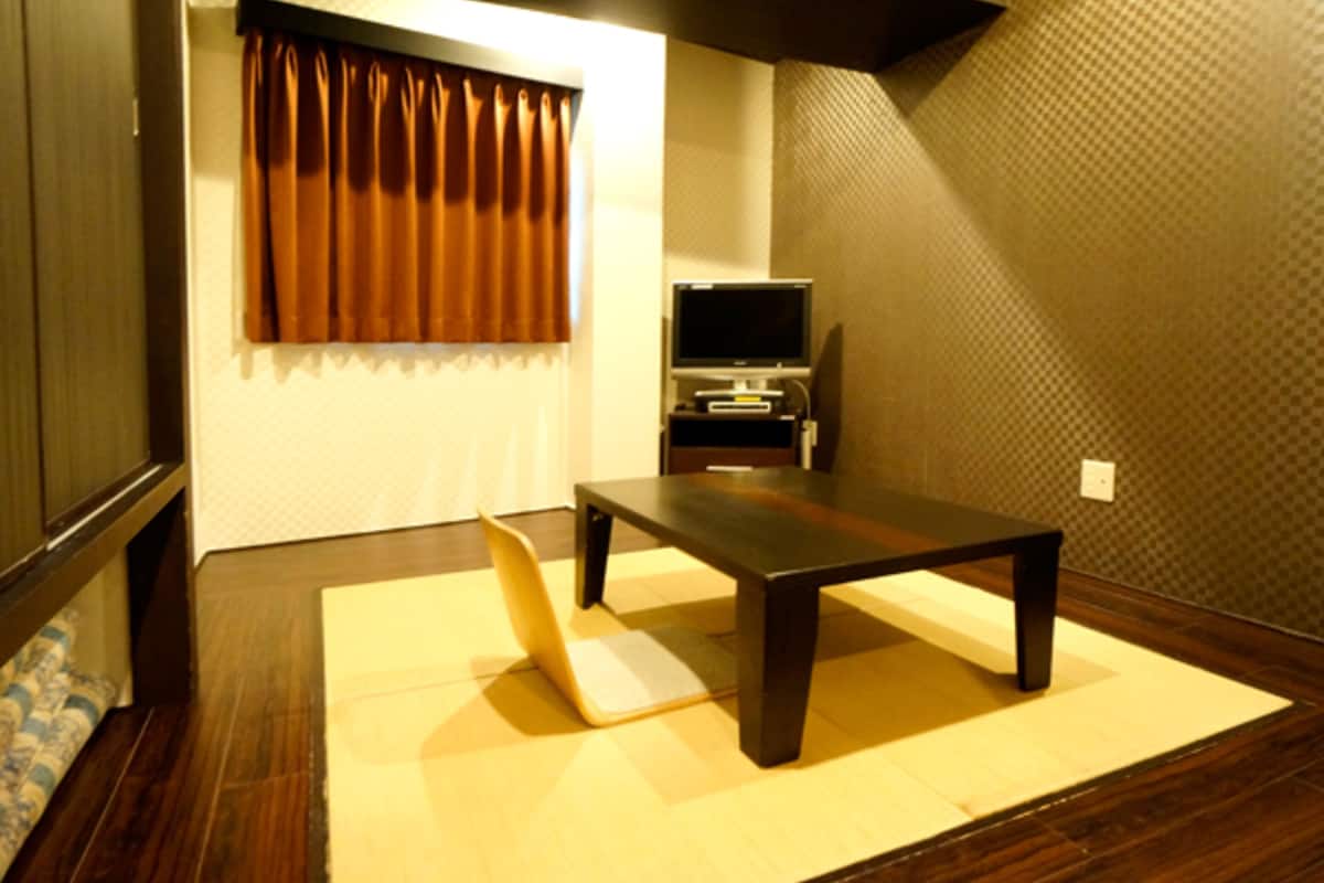 A room with a tatami mat, a dark wooden table, a television on a stand, and a window with brown curtains.
