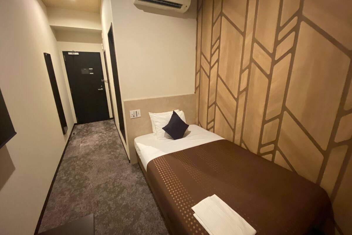 A hotel room with a single bed, a wall-mounted air conditioner, a door, and a carpeted floor.