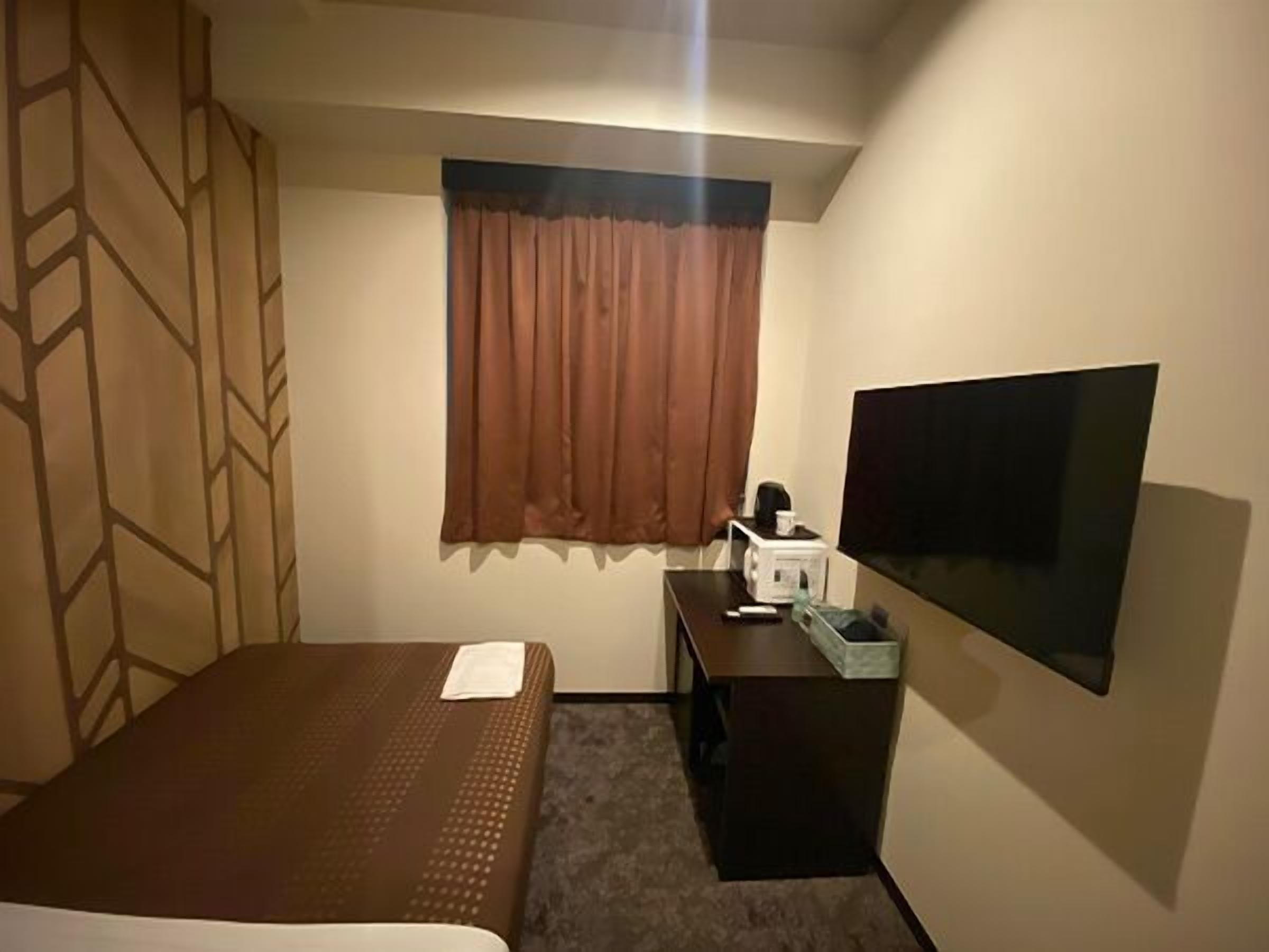 A hotel room with a bed, a desk with a TV, a window with curtains, and a wall-mounted TV.