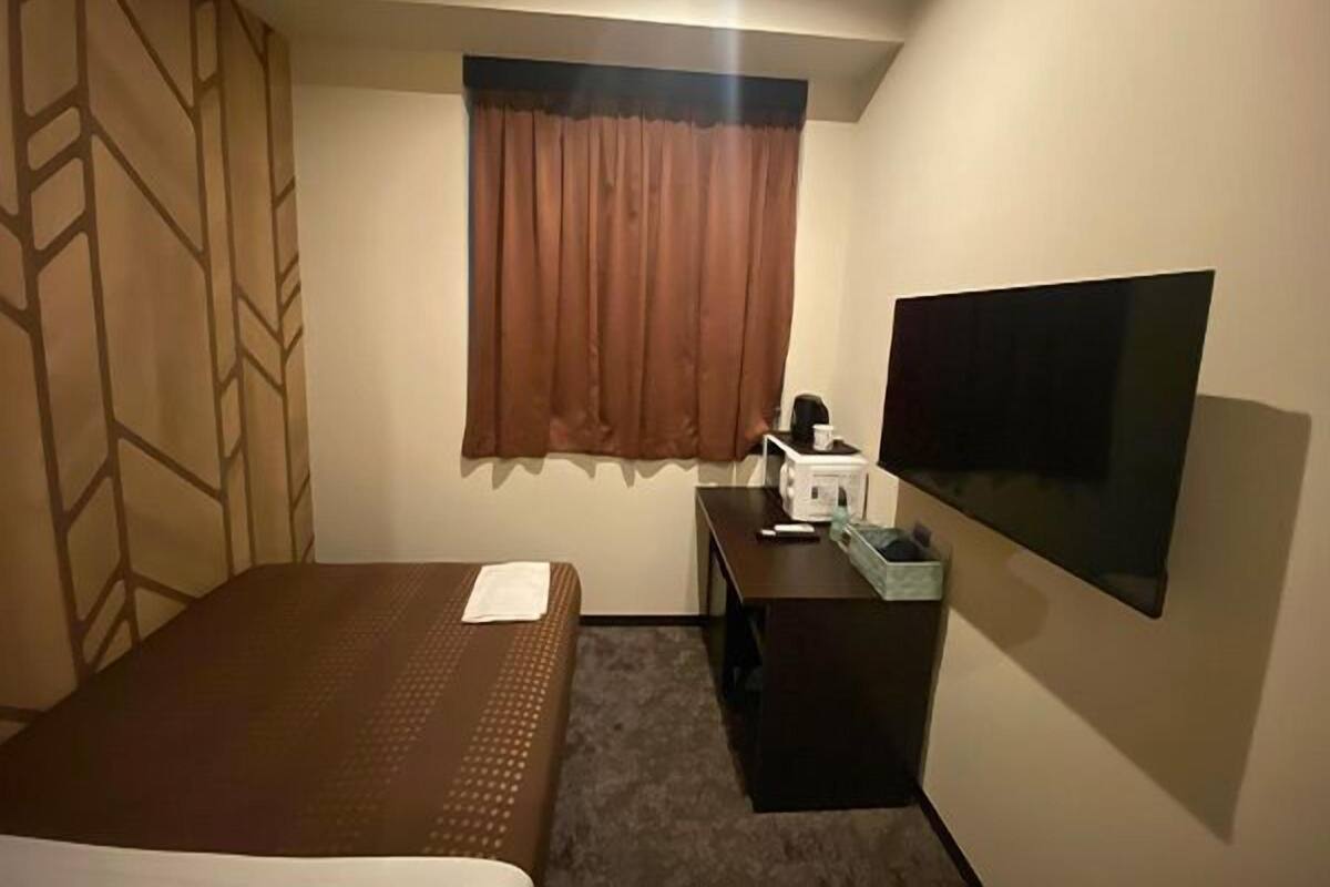 A hotel room with a bed, a desk with a TV, a window with curtains, and a wall-mounted TV.
