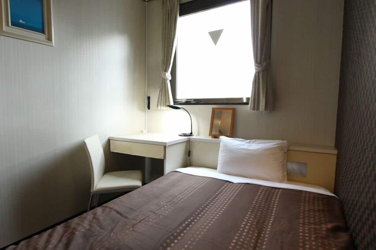 A hotel room with a bed, a desk, a chair, a window with curtains, and a wall-mounted lamp.
