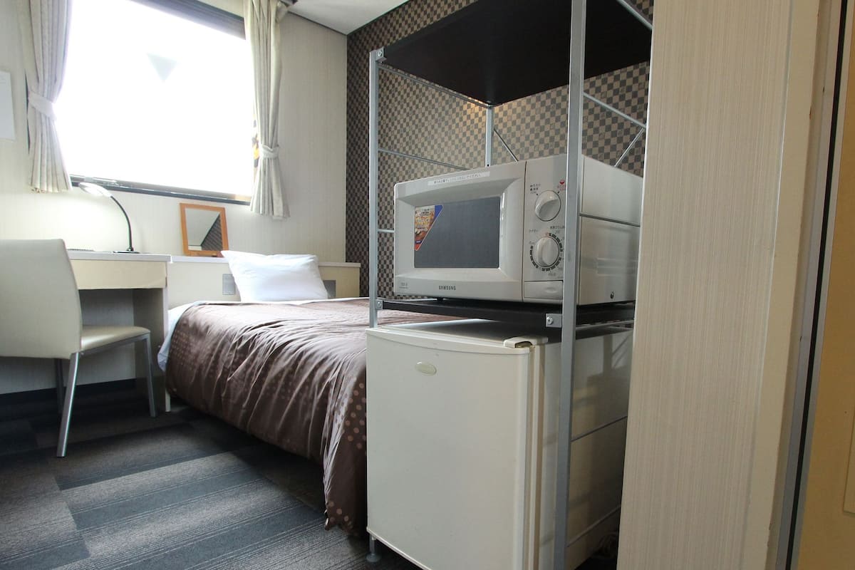 A hotel room with a bed, a desk, a chair, a microwave, and a refrigerator.
