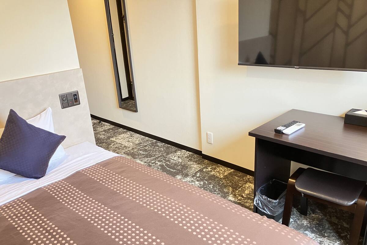 A hotel room with a bed, a desk, a chair, a TV, and a trash bin.