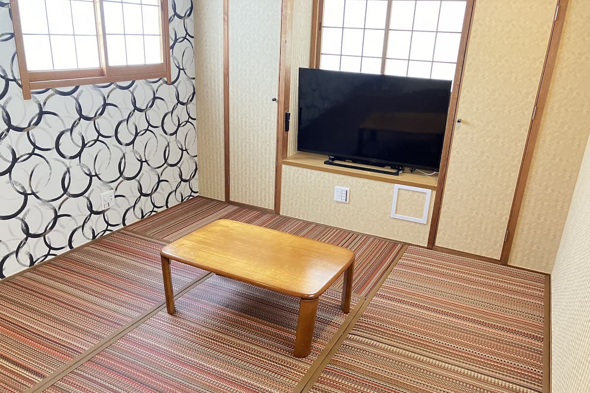 A room with tatami flooring, a wooden bench, a television on a low shelf, and a window with a grid pattern.