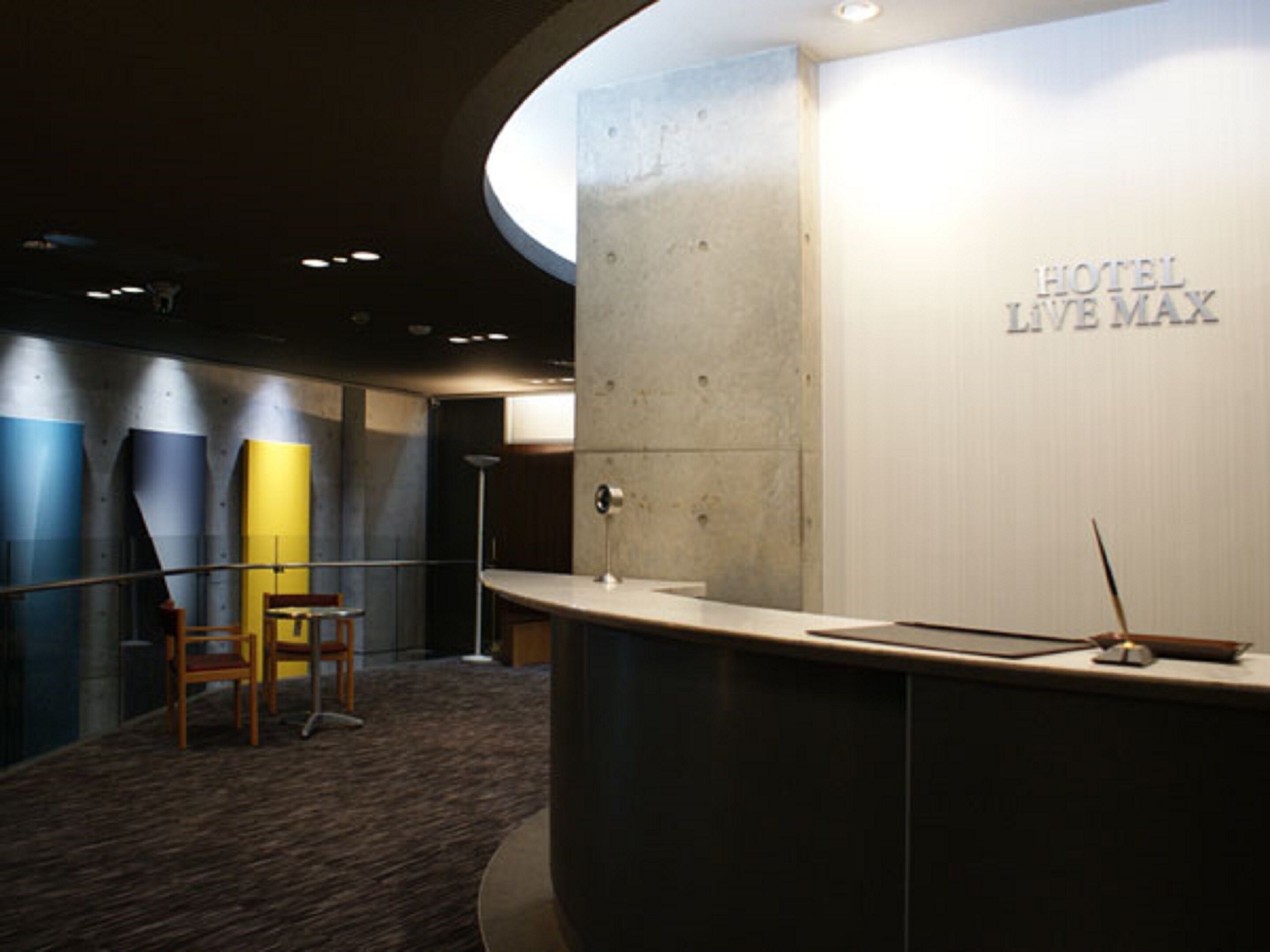 A hotel reception area with a curved counter, a reception desk, and a wall with the hotel's name. There are two chairs and a floor lamp.