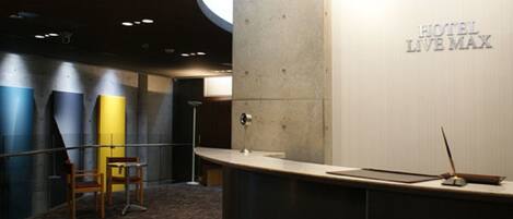 A hotel reception area with a curved counter, a reception desk, and a wall with the hotel's name. There are two chairs and a floor lamp.