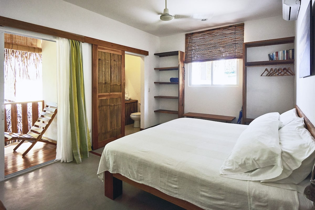 A bedroom with a bed, a wooden chair, a toilet, and a window with blinds.