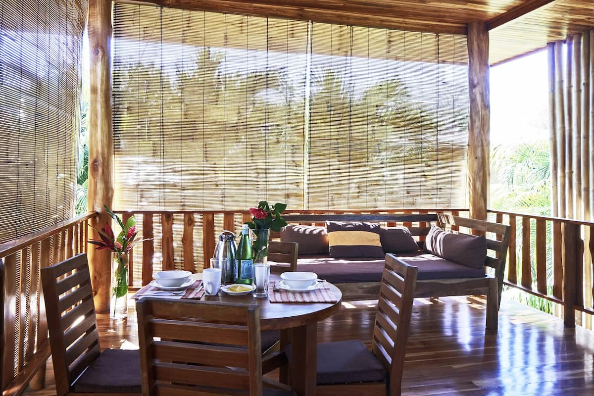 A wooden deck with a table set for dining, a cushioned seating area, and a view of greenery through bamboo blinds.