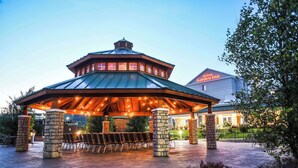 A covered outdoor seating area with stone pillars, a high-roofed pavilion, and a view of the Hilton Garden Inn hotel in the background. - Hilton Garden Inn Watertown/Thousand Islands (Watertown)
