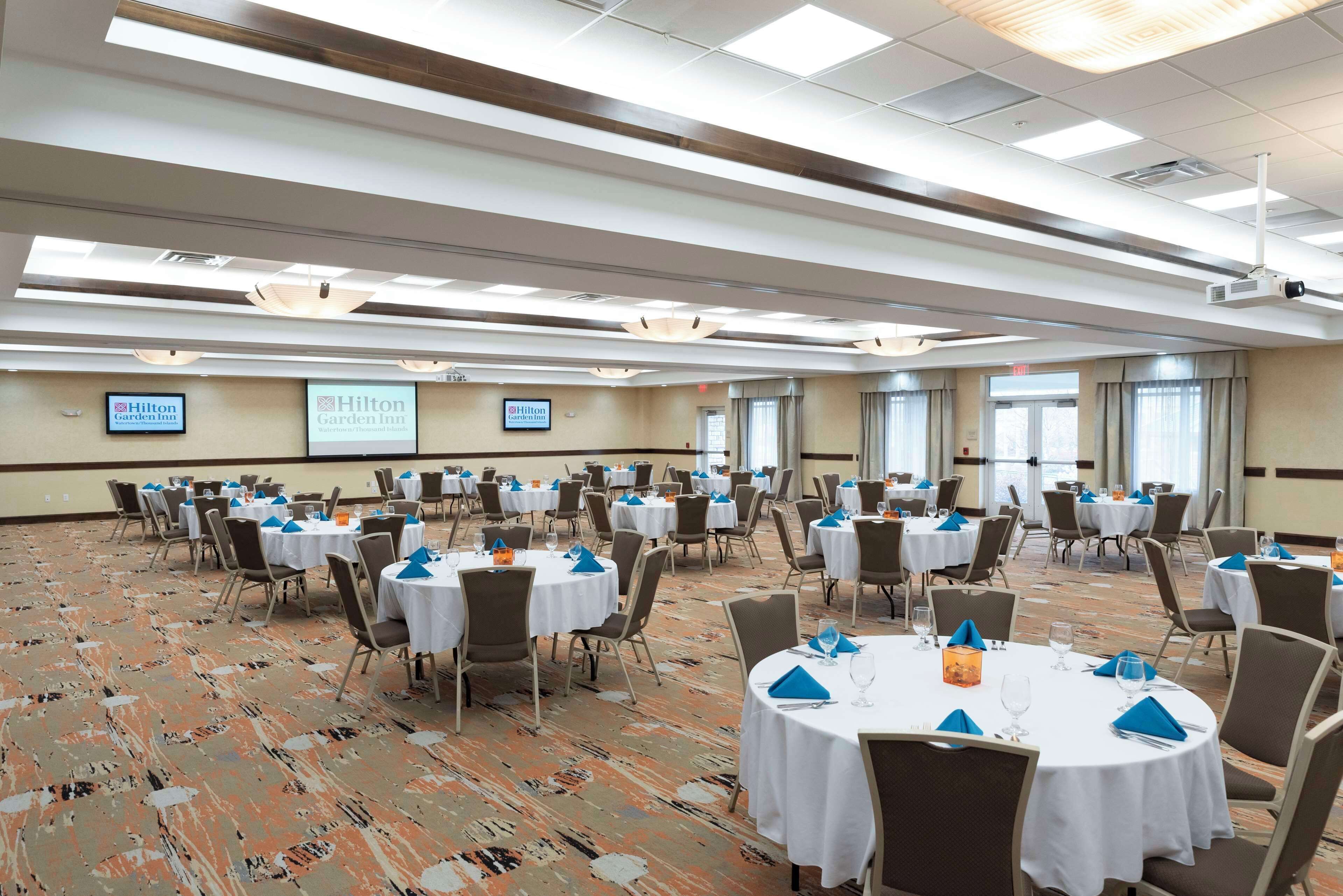 A large conference room with multiple round tables, each set with white tablecloths, blue napkins, and glassware. The room has a carpeted floor and is well-lit with ceiling lights.
