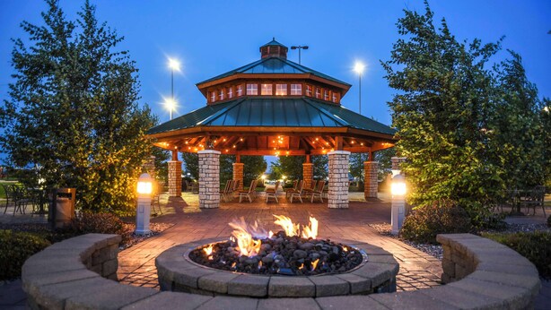 A gazebo with a stone fire pit in the center, surrounded by outdoor seating and a brick pathway. - Hilton Garden Inn Watertown/Thousand Islands (Watertown)