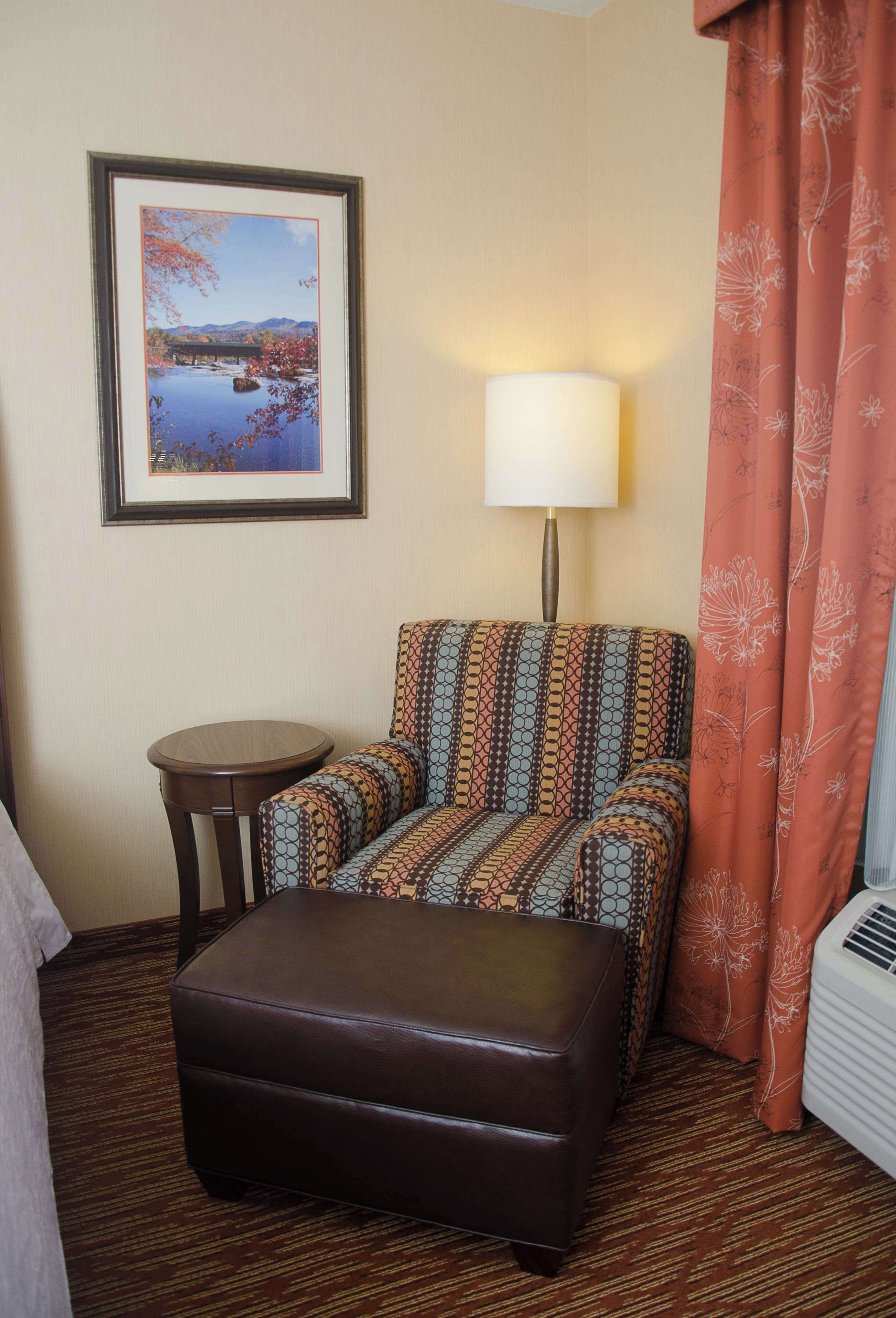 A hotel room with a patterned armchair, a small round table, a brown ottoman, a framed picture on the wall, a lamp, and a window with a curtain.