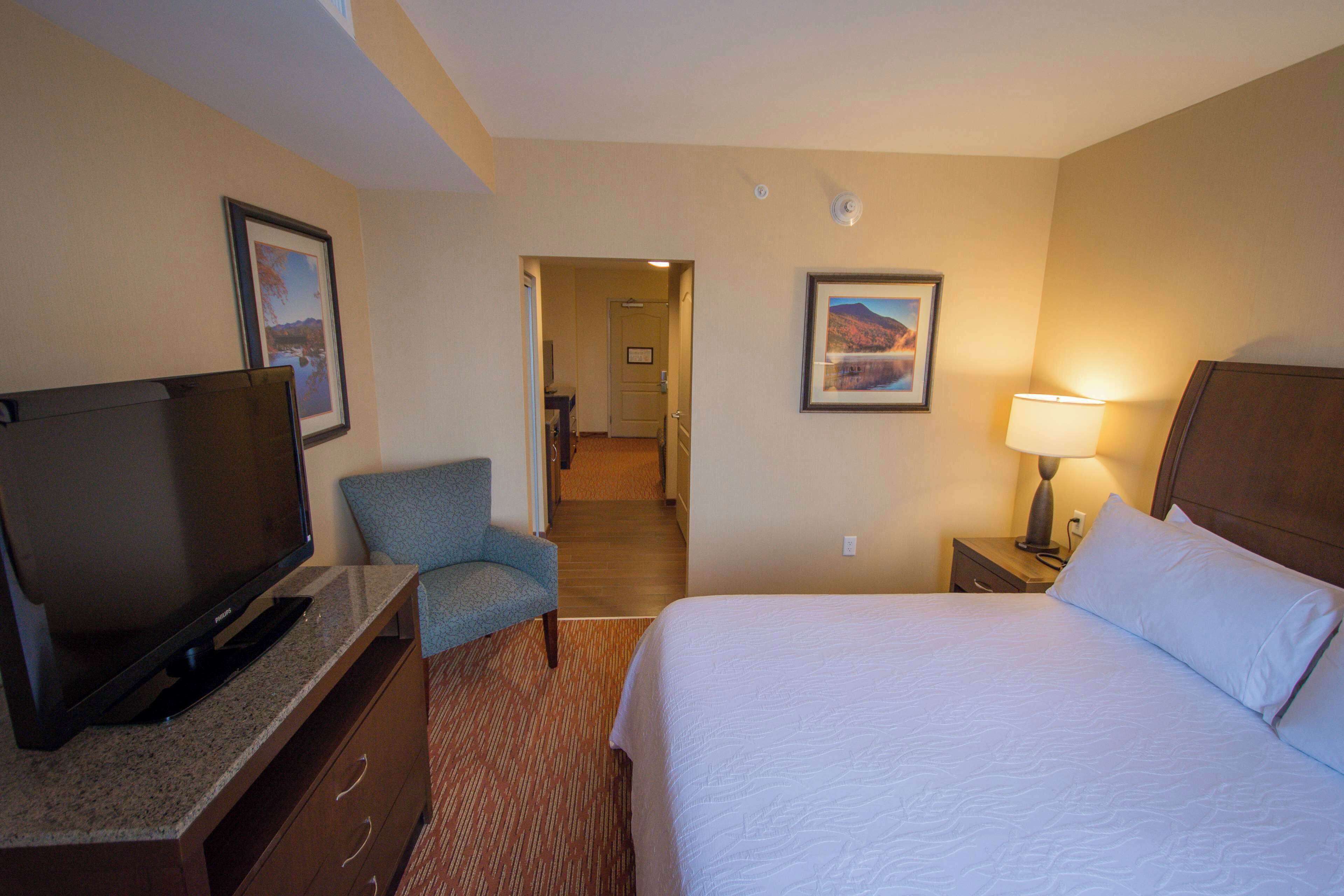 A hotel room with a bed, a television, a chair, a nightstand with a lamp, and framed pictures on the walls.