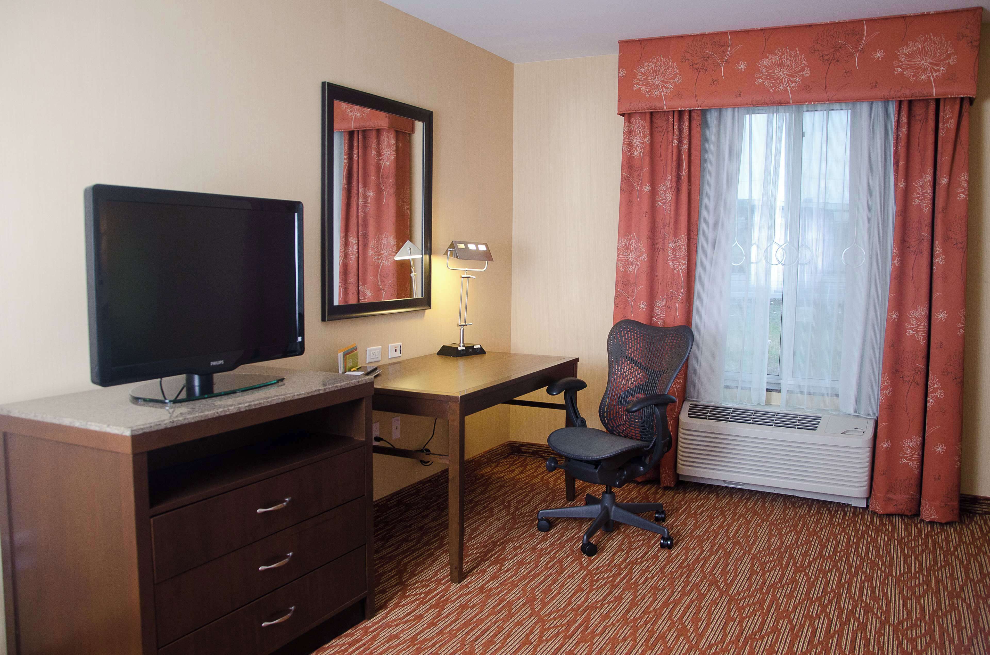 A hotel room with a desk, chair, television, mirror, lamp, and air conditioning unit.