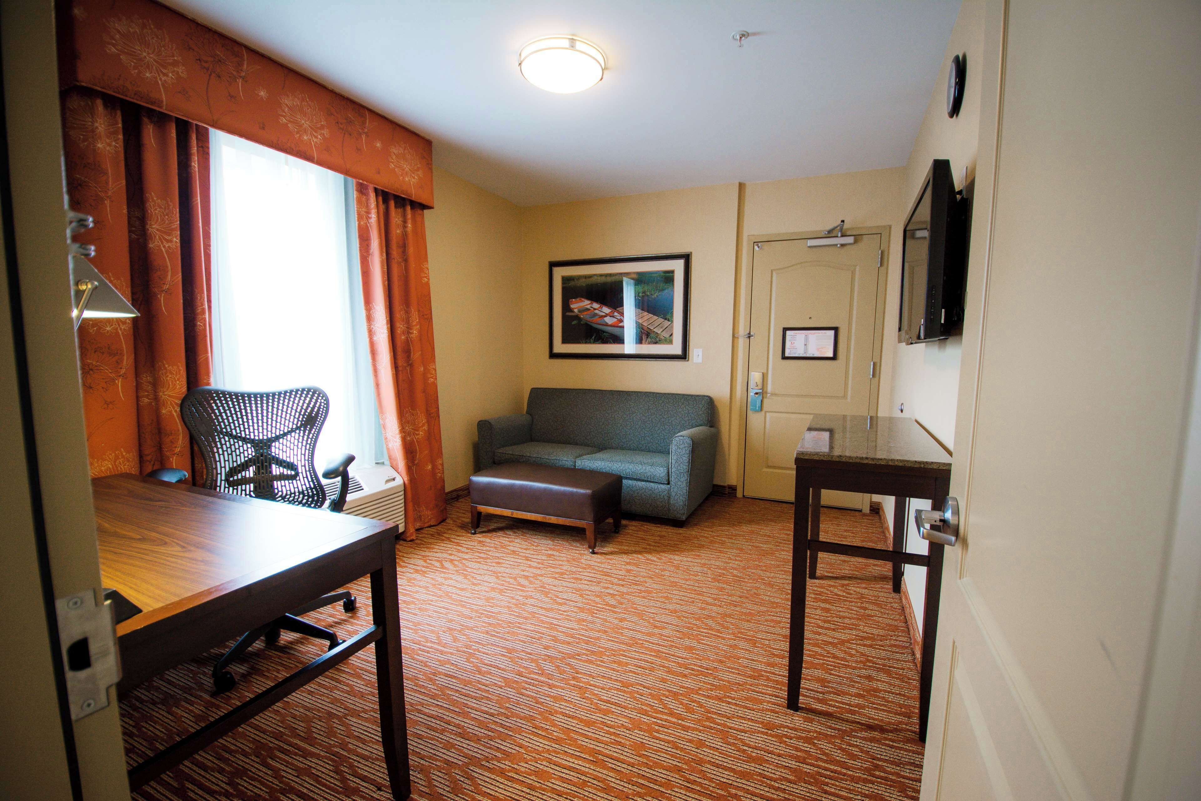 A hotel room with a wooden desk, a grey sofa, a small ottoman, a black office chair, a framed picture on the wall, and a television mounted on the wall.