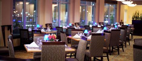 Une salle à manger d’hôtel avec de nombreuses tables et chaises, de grandes fenêtres et un sol recouvert de moquette.