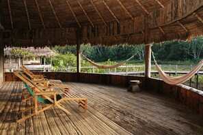 Una terraza de madera con sillones de descanso y una hamaca, rodeada por una estructura de techo de paja y abundante vegetación.