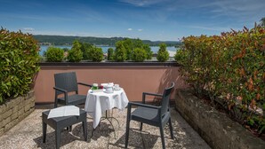 A terrace with two wicker chairs and a table, overlooking a lake with trees and buildings in the background. - Sirio Hotel (Dormelletto)