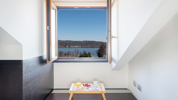 A room with a view of a lake and hills, a small table with breakfast items, and a window with an open shutter. - Sirio Hotel (Dormelletto)
