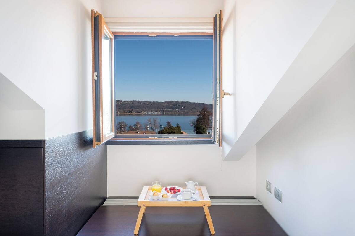 A room with a view of a lake and hills, a small table with breakfast items, and a window with an open shutter.